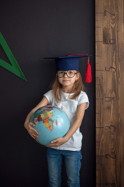 Girl in academic hat and rounded glasses stands at the black wall holding globe