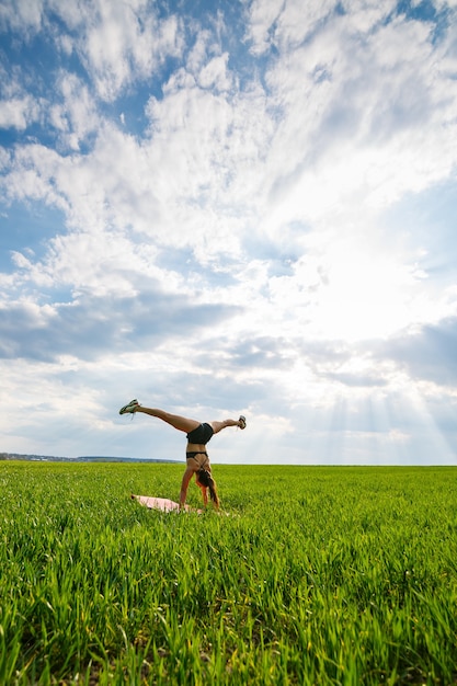 Girl acrobat performs a handstand. the model stands on her\
hands, doing gymnastic splits against the background of green trap\
and blue sky, playing sports in nature