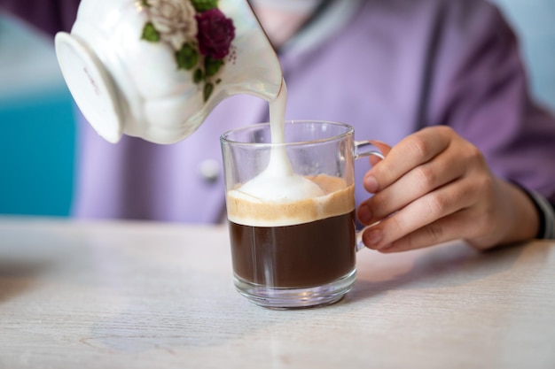 Girl adding milk into glass of delicious coffee with whipped cream