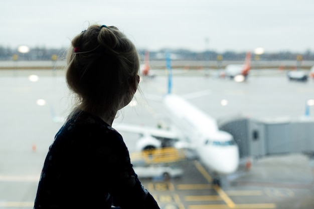 窓の外の空港にいる女の子が飛行機を見て、飛行機を待っている