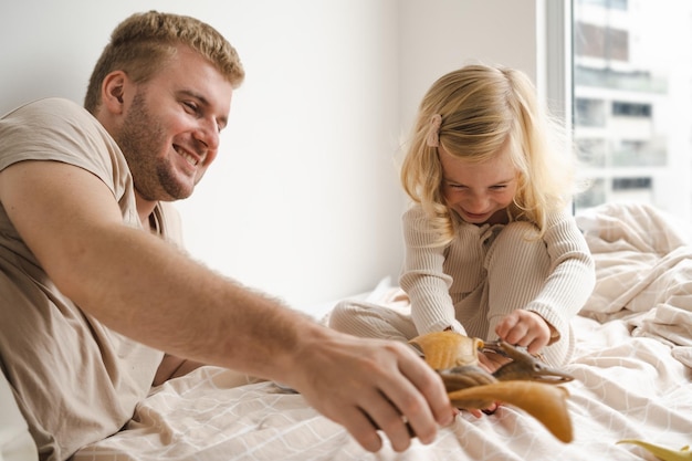 写真 家の窓の近くのベッドでおもちゃで遊ぶ女の子と男ディナサウルス親子が楽しんで峠