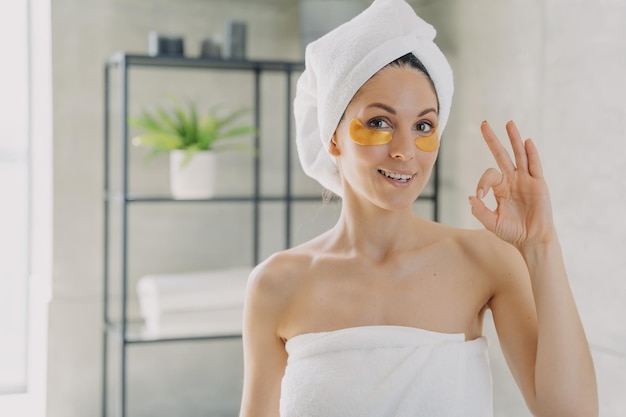 Girl applies collagen eye patches and gestures ok Woman wrapped in towel after spa procedures