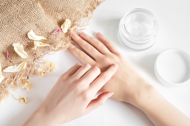 Girl applies moisturizer to her hands on a white wall with dry flower petals and burlap. Ecological natural fragrance-free cream. Female hands with a jar of cream on a light wall