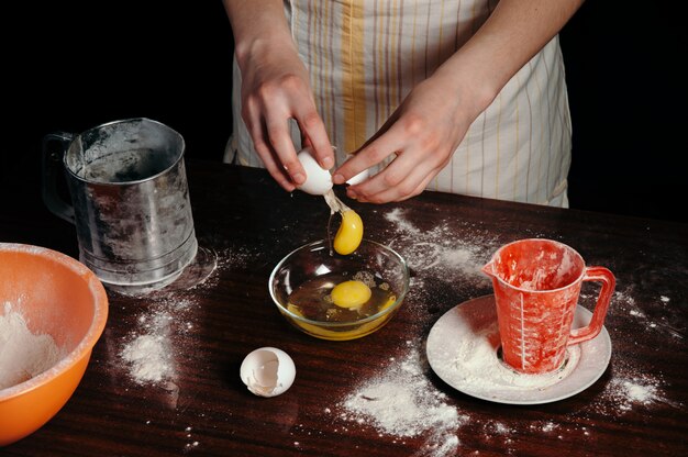 Girl in apron in dark kitchen breaks egg into bowl. 