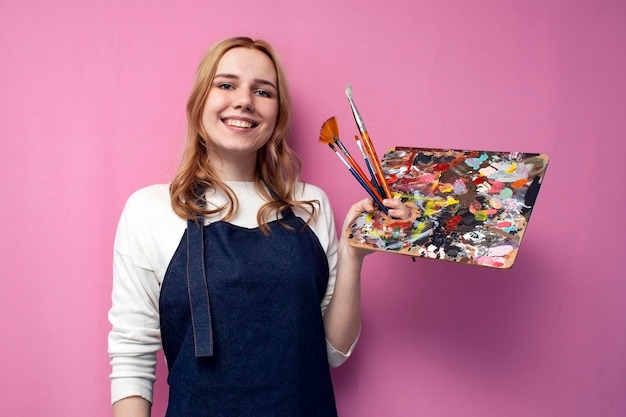 Girl artist holds brushes and a palette and smiles on a pink background