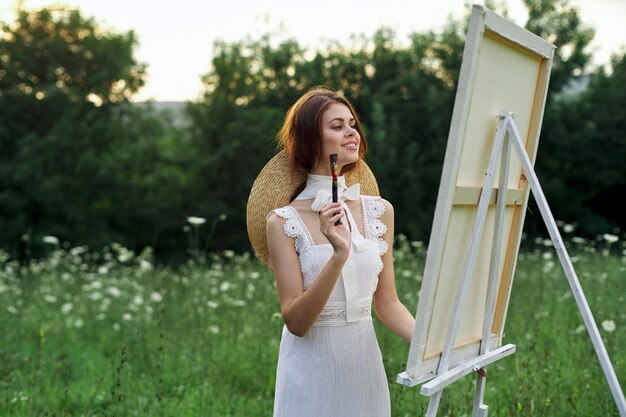 A girl artist in nature with a palette in her hands draws a picture in a beautiful hat