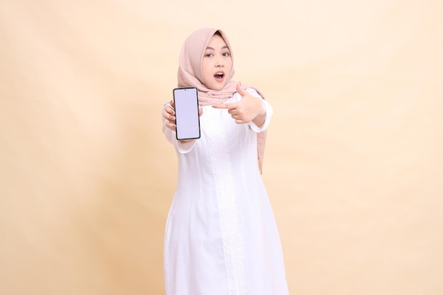 girl asian muslim wearing a hijab in shock holding a cell phone gadget in her hand while pointing at