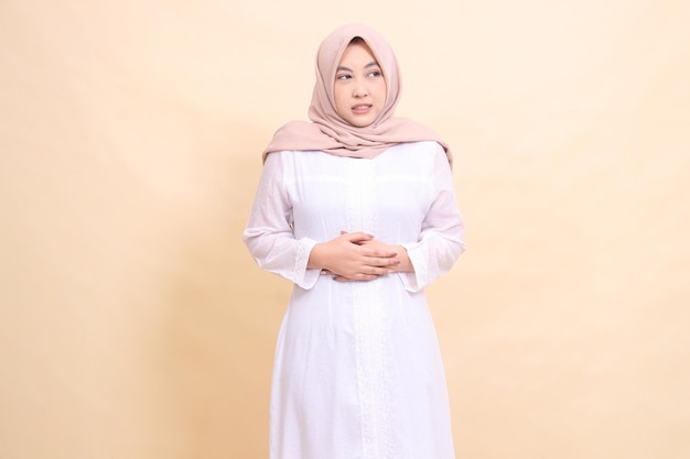 girl asian wearing a white hijab dress stands candidly in pain with both hands holding her stomach m