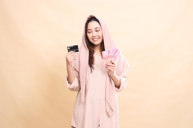 Photo girl asian with candid graceful smile expression hand holding debit card up and carrying rupiah mone