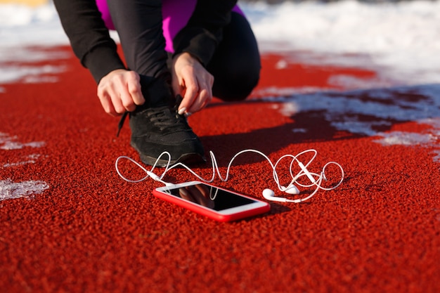 Foto ragazza atleta in scarpe da ginnastica nere, accovacciato sulla pista rossa per correre. vicino c'è un telefono con le cuffie cablate. freddo tempo nevoso