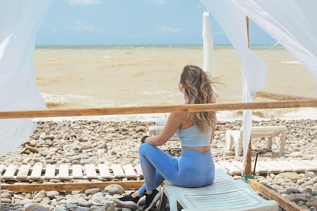 girl athlete in blue sportswear is resting after fitness on the seashore in a beach gazebo