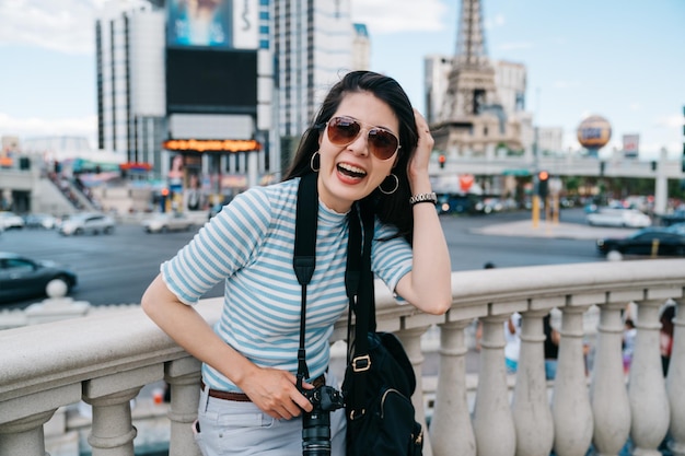 Girl backpacker cheerful looking face camera and smiling\
attractive. charming lady tourist holding vintage camera while join\
sightseeing city tour in las vegas. famous hotel with tall tower in\
back