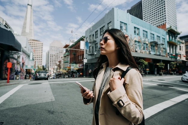 Photo girl backpacker walking on street in chinatown holding mobile phone searching direction online map app on sunny day. transamerica pyramid view tall tower in city urban san francisco with blue sky.