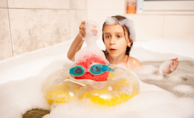 Girl bathes in a bath with foam and plays life-saving ball and swimming goggles
