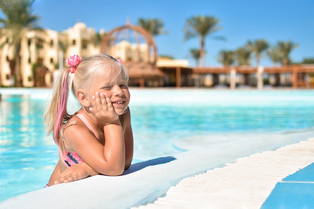 A girl in a bathing suit sits in the azure water of a swimming pool in a hotel with palm trees