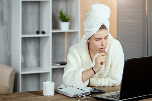 A girl in a bathrobe and a towel on her head, watching movies on a laptop and eating chocolate