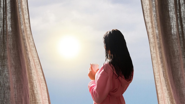 Девушка в халате с мокрыми волосами и чашкой кофе смотрит на восход солнца