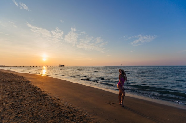 海の近くのビーチで女の子が日没のスタイリッシュな水着とカンカン帽を背景にポーズします。