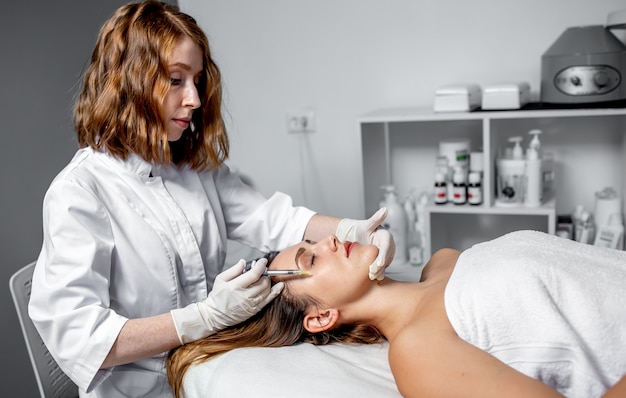 Girl beautician makes injections on the lips and face of a beautiful woman in a cosmetic wedge