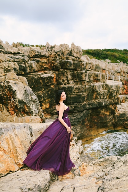 Girl in a beautiful lilac dress fluttering in the wind stands on a rock against the background of