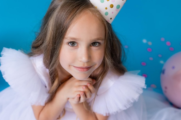 girl in a beautiful white fluffy dress and a festive hat is sitting on the floor on blue background