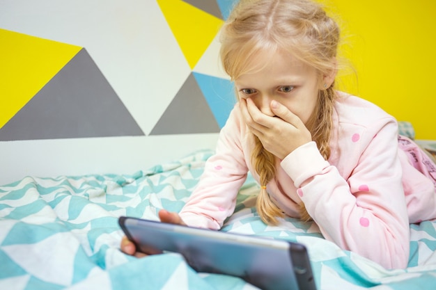 Girl on the bed in the nursery play on the tablet