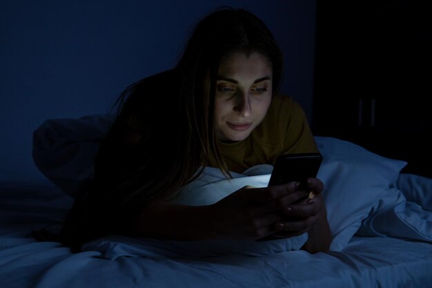 A girl in bed with a phone on a white bed in a dark room