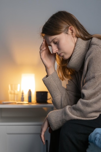 Girl in beige sweater sitting on bed and holding her head Woman suffering from headache or migraine