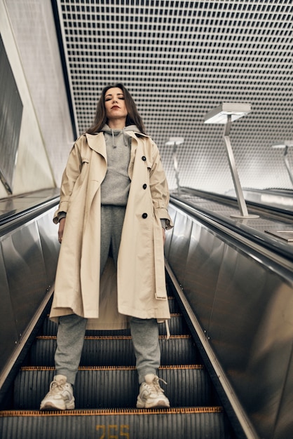 A girl in a beige trench coat descends the subway on an escalator.