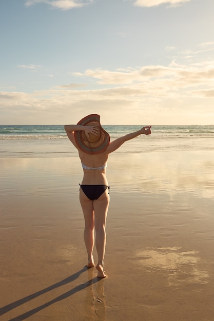 Foto ragazza in bikini con cappello di paglia sulla riva di una spiaggia che tiene il suo cappello di paglia.