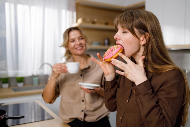 Photo girl bites an appetizing donut while having tea with her mom mother and daughter in the kitchen