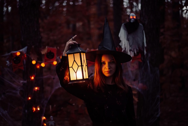 Girl in black clothes witch hat standing with lantern in dark forest Concept of witchcraft halloween