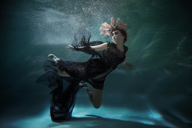 A girl in a black dress poses under water against a dark background, as if she is in weightlessness