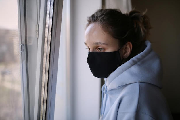 Photo the girl in black mask is standing at the window of the house in isolation