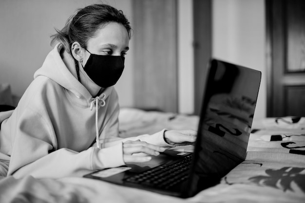 Girl in a black mask works on a laptop at home in isolation black and white photo