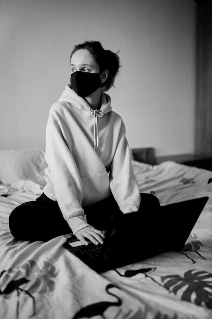 Girl in a black mask works on a laptop at home in isolation black and white photo