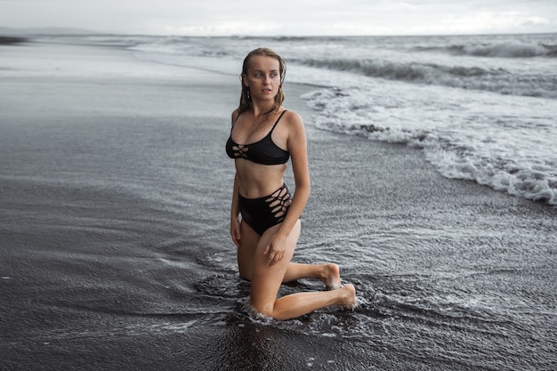 The girl in the black swimsuit on the black sand is kneeling