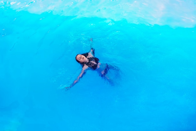 Girl in black swimsuit and sunglasses swims on her back in clear water in the pool beautiful woman i