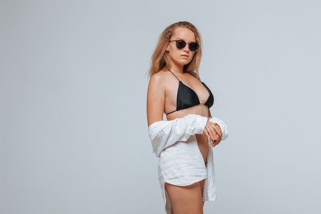 Girl in a black swimsuit and sunglasses and a white shirt on a light gray background