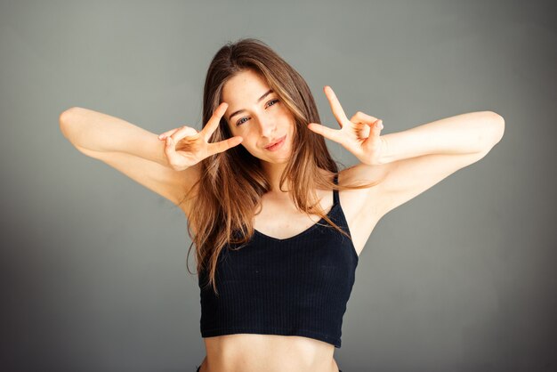 A girl in a black top with her hair showing both hands shows a victory sign. Two fingers. Number two. On a gray wall. Without makeup, without retouching.