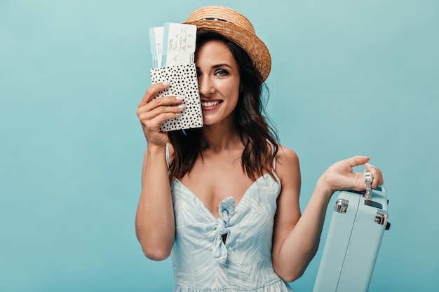 Girl in blue dress covers her face with passport tickets and holds suitcase