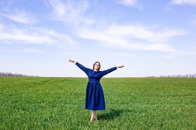 A girl in a blue dress stands in a green field of wheat. Spring landscape. Agriculture.