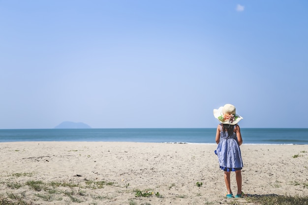 青いスカートと海のバックグラウンドを持つ海岸に青いドレスと帽子の立っている服を着た女の子