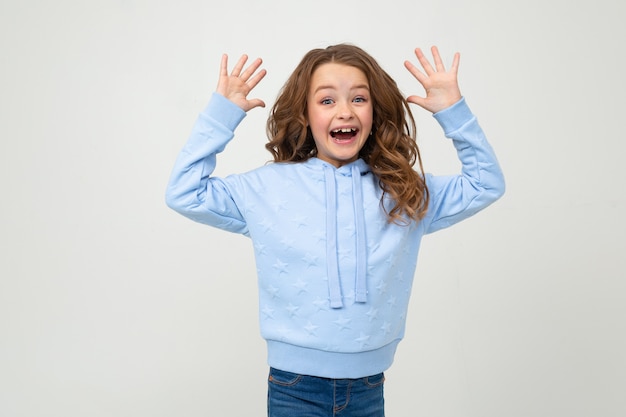  girl in a blue hoodie smiles joyfully while holding her palms in front of her on a light gray wall