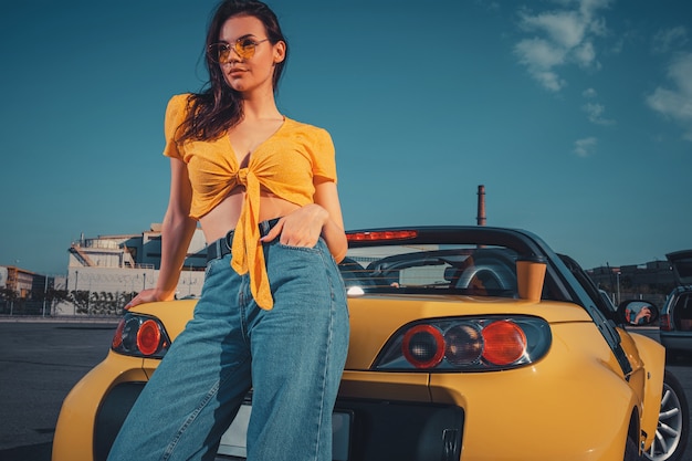 Girl in blue jeans orange top and sunglasses is posing leaning on yellow car cabriolet with paper cup of beverage on trunk at parking lot