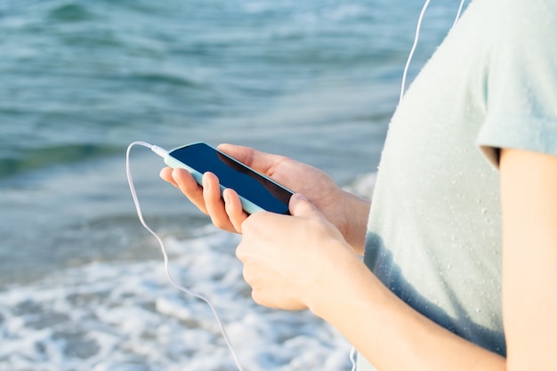 Girl in a blue shirt holding a mobile phone in hand and listening to music with headphones on the beach 