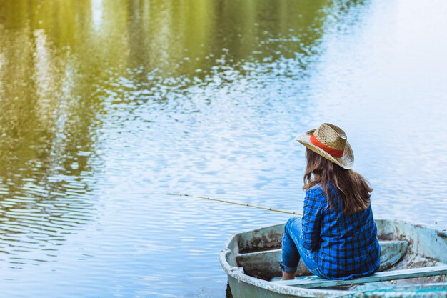 青いシャツを着た女の子が座って、夏の麦わら帽子の湖でボートに乗って釣りをしています