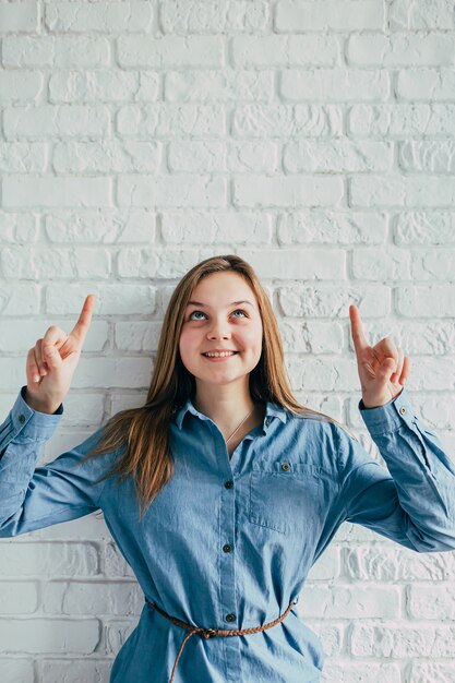 Foto ragazza in una camicia blu con le mani in su contro il muro di mattoni bianco