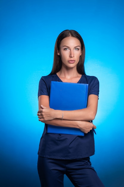 Girl in a blue surgical suit posing standing on a blue background hugging a blue tablet for records in her hands and a pen.Care for people.Medicine.