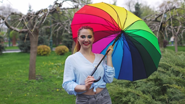 Girl in a bluish shirt with bright makeup and long colored braids. Holding an umbrella in the colors of the rainbow on a flowered park enjoying the coming spring